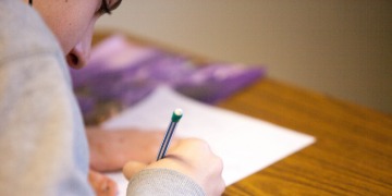 Das Bild zeigt links im Bildrand das Gesicht und die rechte Schulter einer weibliche Person. Sie hält einen Stift und der rechten Hand und schreibt konzentriert auf ein Blatt. Nach rechts hin ist das verschwommen. 